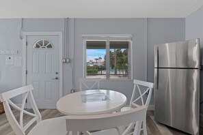 Eine moderne Küche mit einem Edelstahlkühlschrank, einem weißen Esstisch mit Stühlen, einer weißen Tür mit Fenster und Blick auf einen Pool sowie Palmen im Freien.