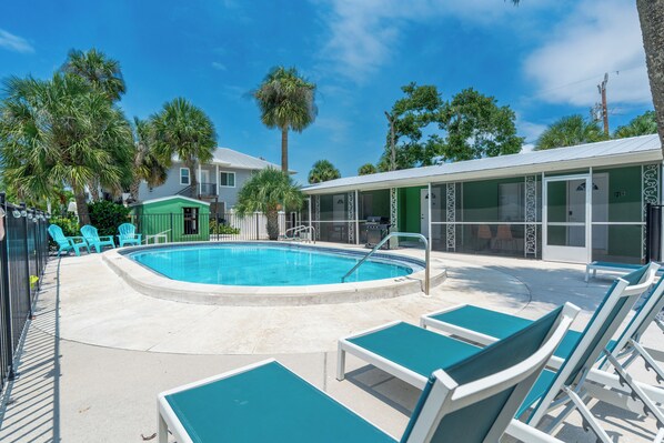 A pool area with lounge chairs, a small building with a green exterior, and palm trees. - 🐾 Dune Your Thing B | Pet Friendly FMB | Heated Pool + Beach Access (Fort Myers Beach)