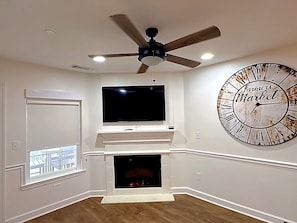 A living room with a fireplace, a mounted TV, a ceiling fan, and a large clock on the wall.