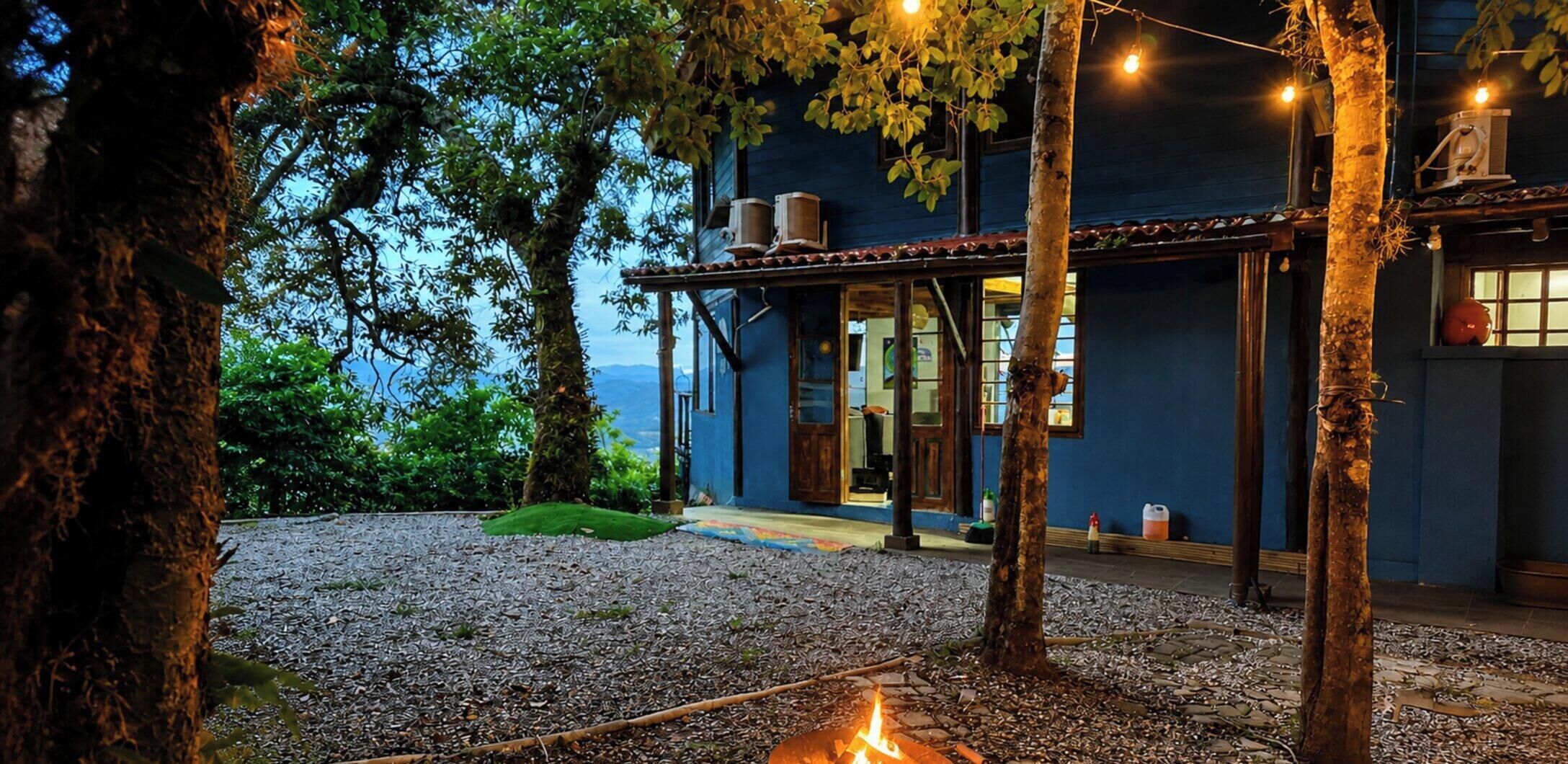 Uma área externa rústica com lareira, árvores e um edifício com fachada azul.