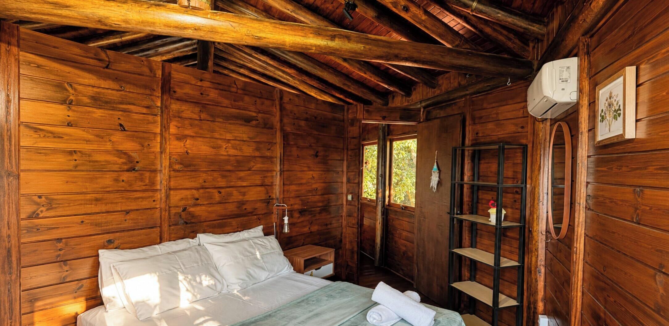 A wooden cabin room with a bed, a shelf, and a window.