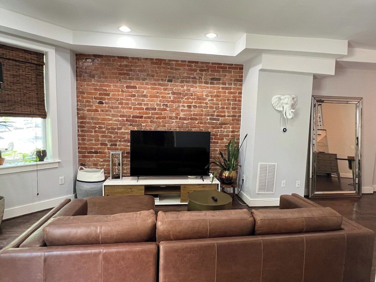 Un salon avec un mur en briques, un grand téléviseur à écran plat, un canapé en cuir marron et une petite table d’appoint.