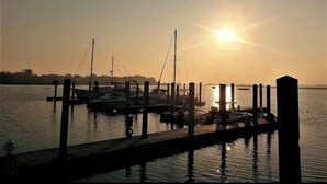 Eine Marina mit vertäuten Booten, eine Pier, die sich ins ruhige Wasser erstreckt, und eine untergehende Sonne im Hintergrund.