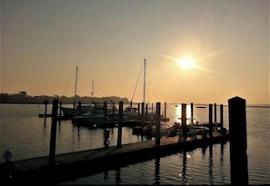 Eine Marina mit vertäuten Booten, eine Pier, die sich ins ruhige Wasser erstreckt, und eine untergehende Sonne im Hintergrund.