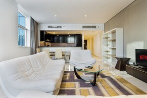 Ein modernes Wohnzimmer mit Sofa, Sessel und Couchtisch, im Hintergrund eine Küche und ein Fernseher auf einem Schrank.