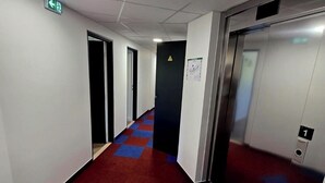 Een hotelgang met een rood-blauw geruit tapijt, een lift en een deur met een waarschuwingsbord.