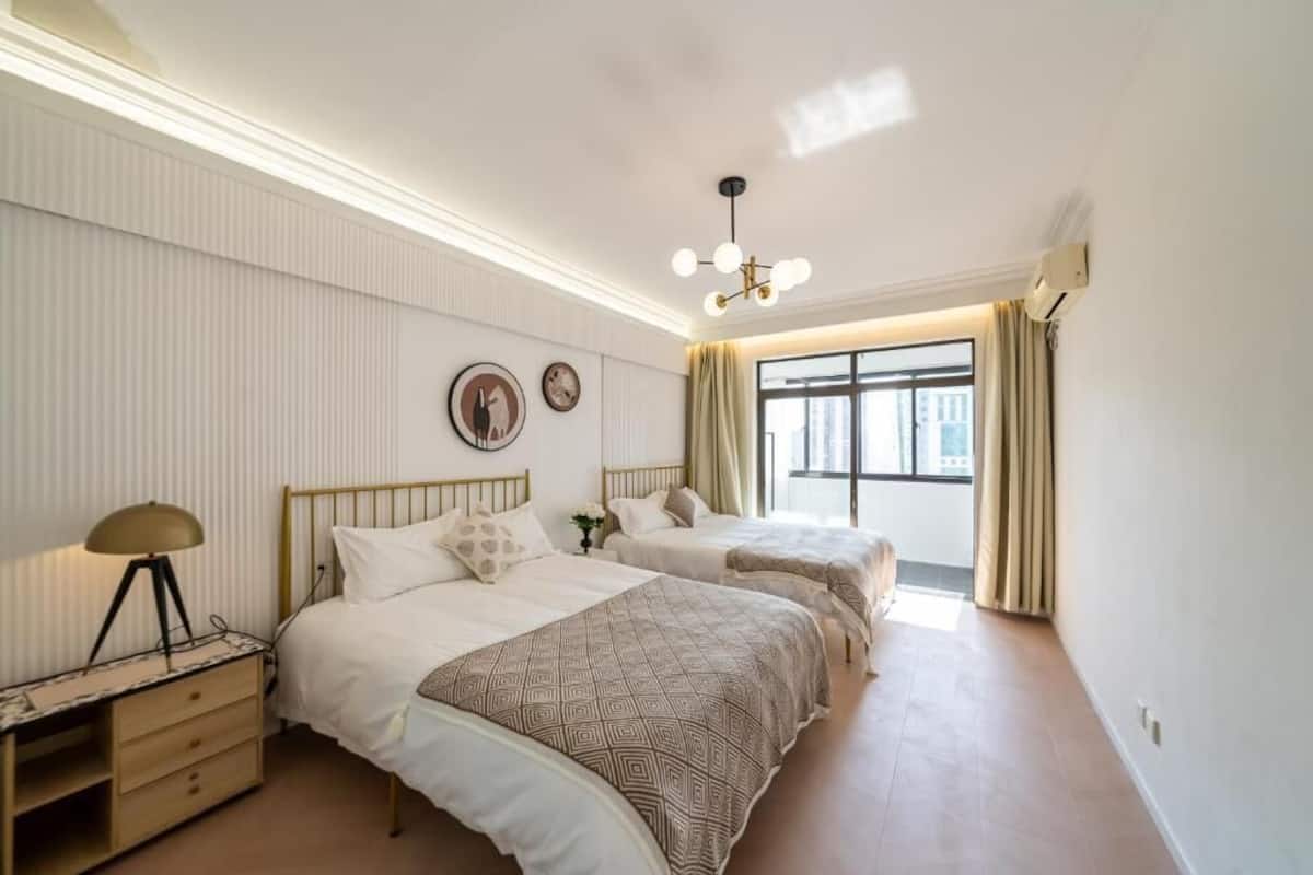 Une chambre avec deux lits, une table de chevet, une lampe, un luminaire au plafond et une fenêtre avec des rideaux.