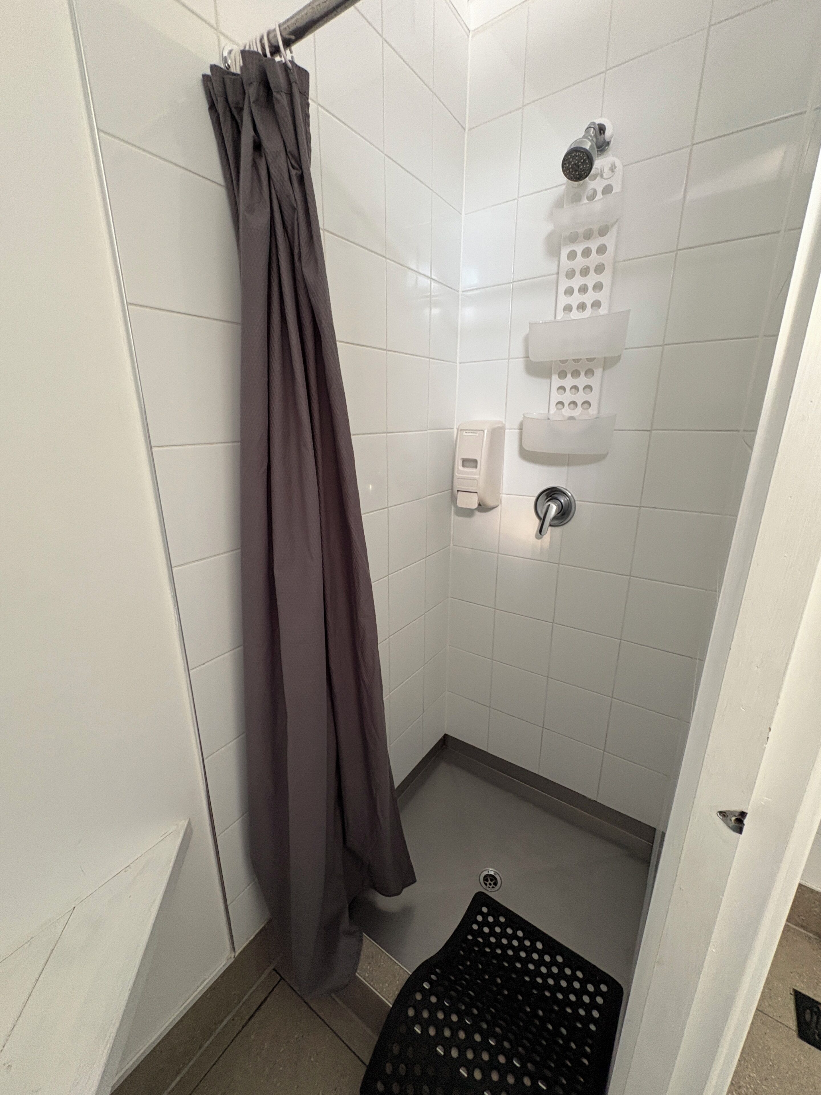 Un espacio de ducha pequeño con azulejos, un cabezal de ducha, dispensador de jabón y una alfombra negra.