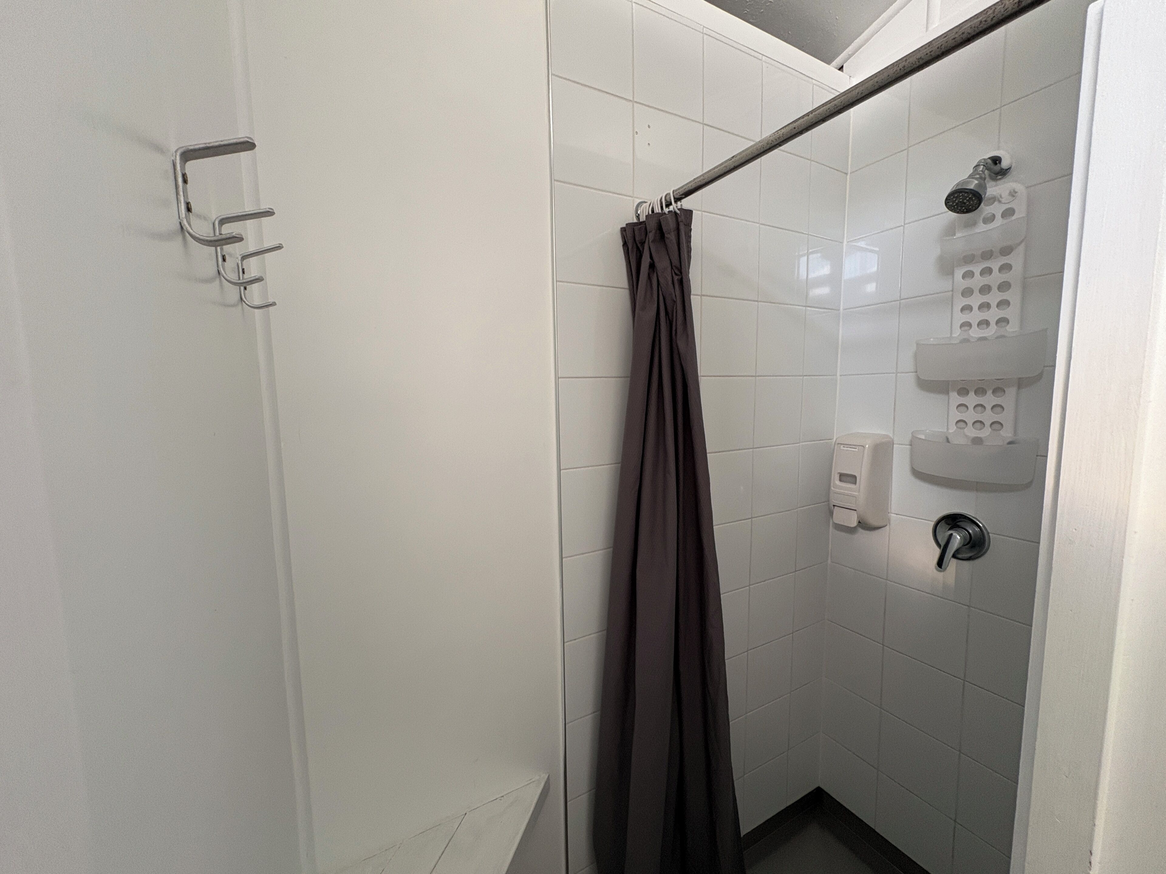 A small, white-tiled shower stall with a showerhead, a handheld showerhead, and a soap dispenser.