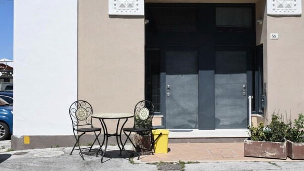 Un edificio con una puerta negra grande, una mesa pequeña con dos sillas y un bote de basura amarillo.