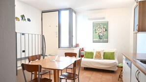 Un espacio habitable compacto con mesa, sillas, sofá, kitchenette y un ventanal pequeño.
