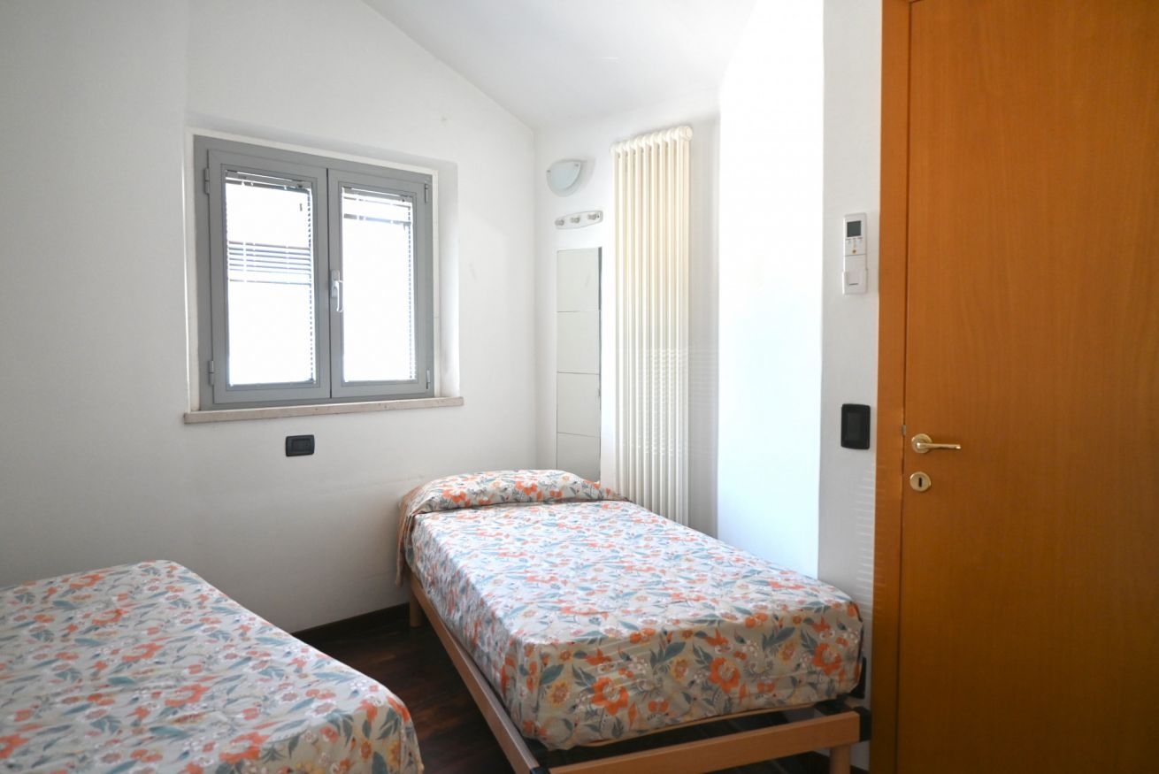 A small, single-room with two beds, a window with blinds, a radiator, and a wooden door.