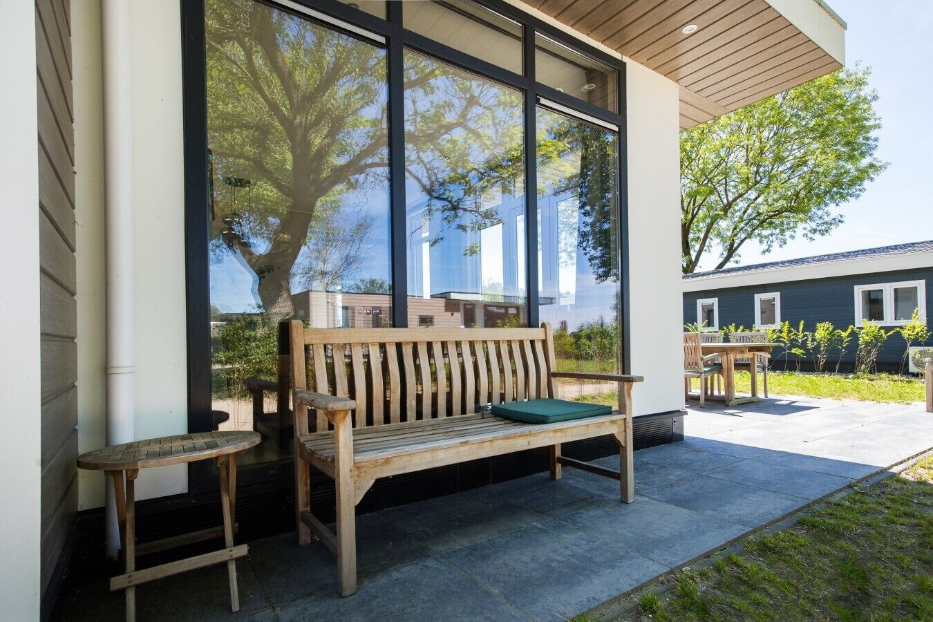 Um banco de madeira com almofada, uma mesa pequena e uma janela grande com vista para árvores e edifícios.