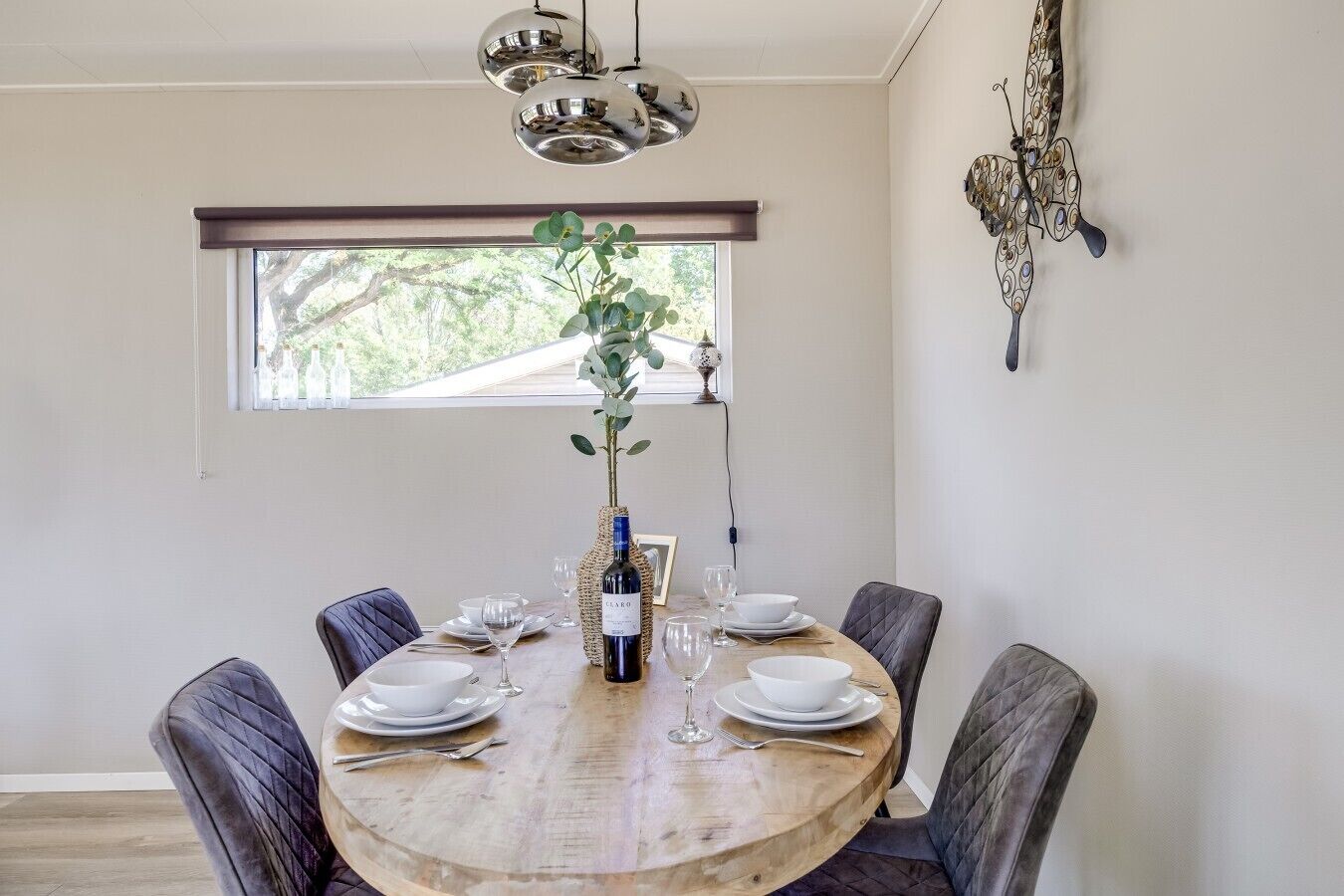 A dining area with a round wooden table, six chairs, a vase with a plant, a bottle of wine, and a decorative wall piece.