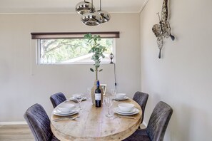 A dining area with a round wooden table, six chairs, a vase with a plant, a bottle of wine, and a decorative wall piece.