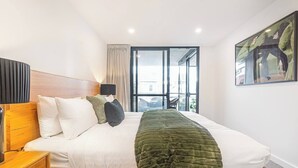 Une chambre d’hôtel avec un lit, une tête de lit en bois, une table de chevet, une lampe, une fenêtre avec des rideaux et un tableau au mur.