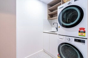 Une buanderie Ă©quipĂ©e de deux machines Ă laver superposĂ©es, dâun petit Ă©vier et dâune Ă©tagĂšre au-dessus des machines.