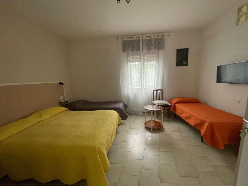 A room with two beds, a small table, a chair, a window with curtains, and a flat-screen TV mounted on the wall.