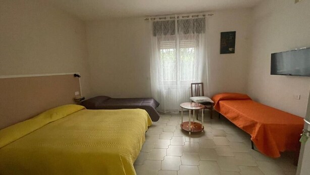 A room with two beds, a small table, a chair, a window with curtains, and a flat-screen TV mounted on the wall. - Mosaico (Pompei)