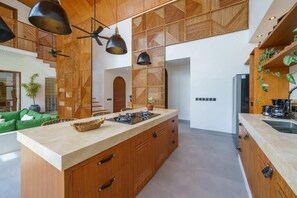 A modern kitchen with a central island, wooden cabinetry, pendant lighting, and a view of the living area. - K Club Greenfields (Badung)