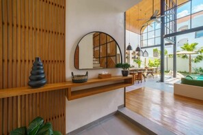 A modern interior with a wooden wall, a round mirror, a shelf with decorative items, and a view of an outdoor area with plants and a pool. - K Club Greenfields (Badung)