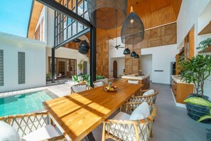 A spacious outdoor area with a wooden dining table, wicker chairs, a pool, and a large window overlooking the pool. - K Club Greenfields (Badung)