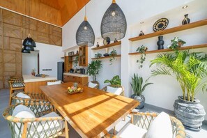A dining area with a wooden table and wicker chairs, surrounded by potted plants and hanging pendant lights. - K Club Greenfields (Badung)