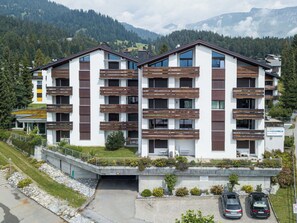 Ein modernes Mehrfamilienhaus mit Balkonen, einem Parkplatz und einer Bergkulisse.