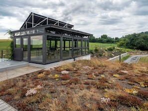 A modern glass structure with a metal roof, surrounded by a natural landscape with diverse vegetation.