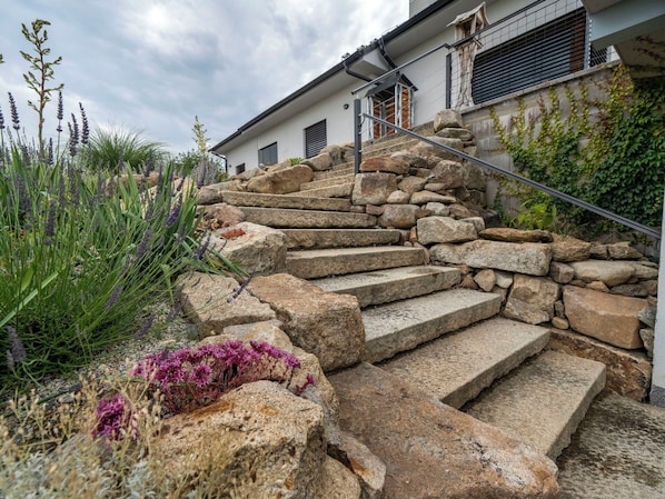 A stone staircase leading to a building with a metal handrail, surrounded by various plants and flowers.