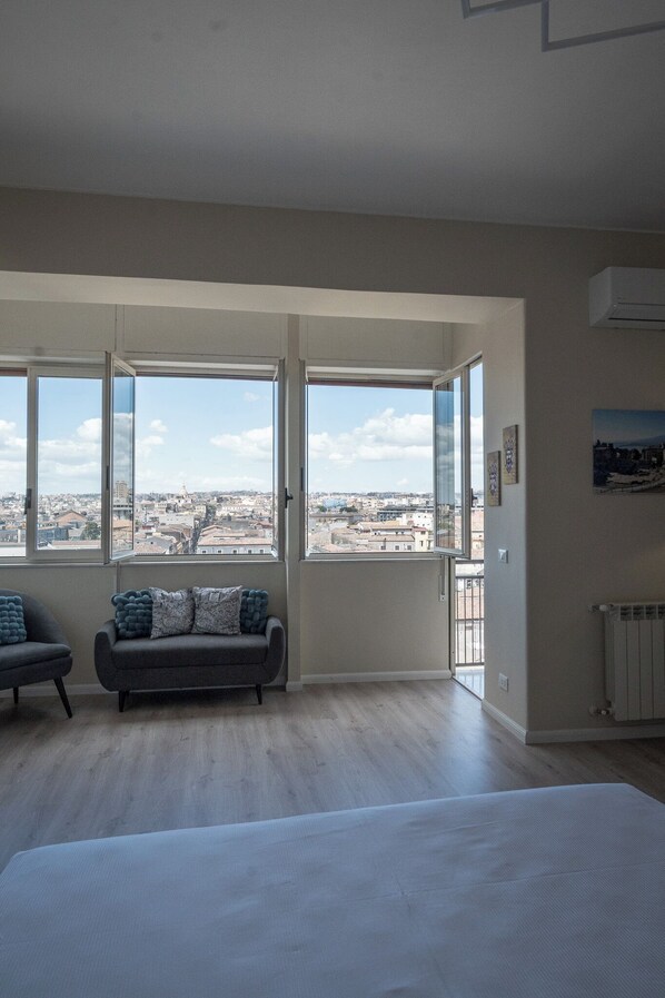 Un dormitorio con una cama grande, un sofá gris con cojines azules y vistas a la ciudad a través de amplias ventanas.