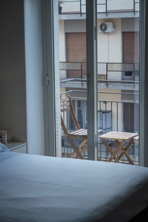 Ein Zimmer mit einem Bett, einem Fenster mit Blick auf einen Balkon mit Stuhl und Tisch sowie einer Klimaanlage.