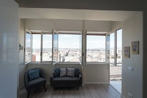 Una habitación con sofá y sillón frente a grandes ventanales con vista a la ciudad.