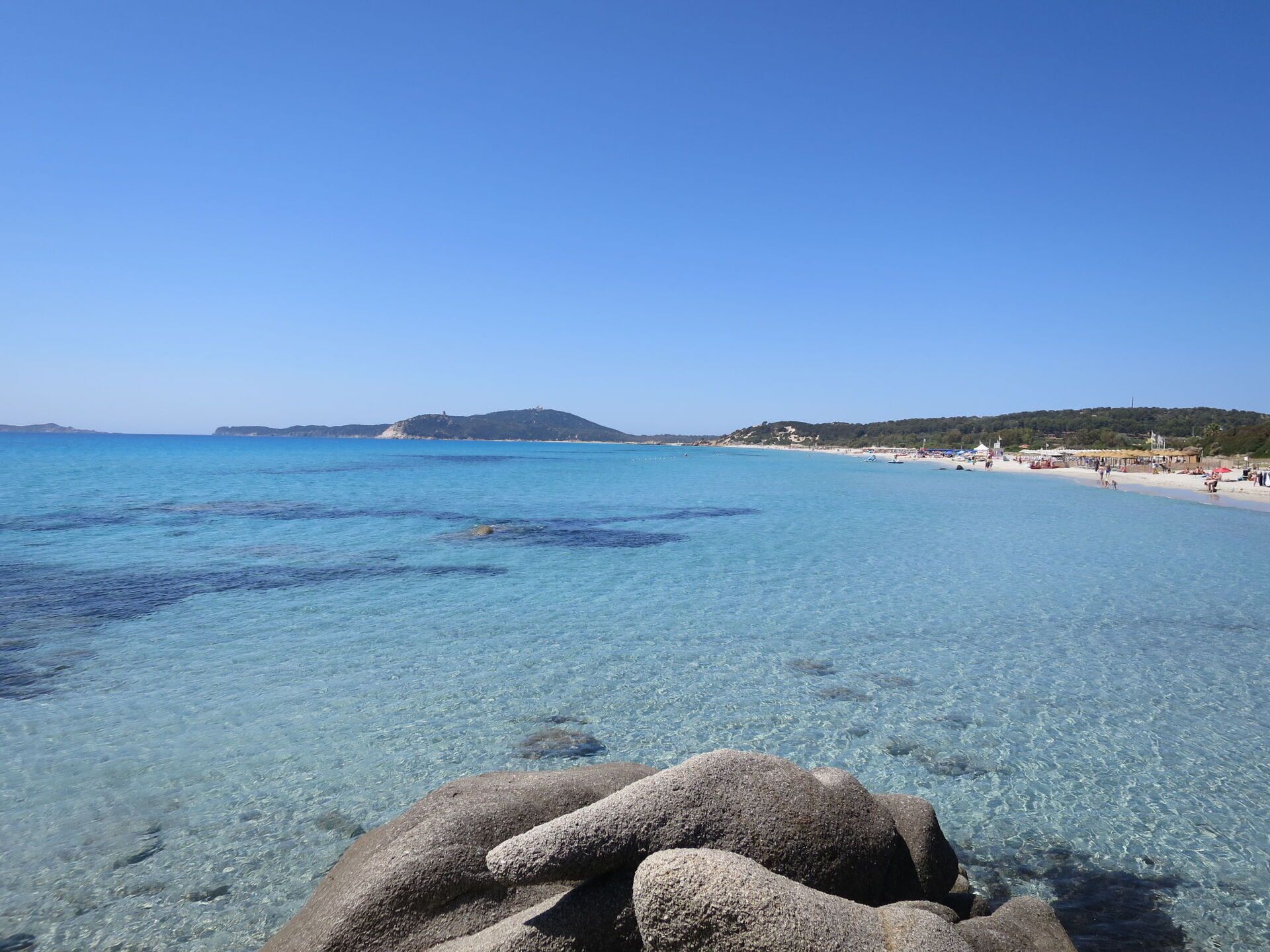 A clear coastal beach with rocky formations in the foreground and a distant hill.