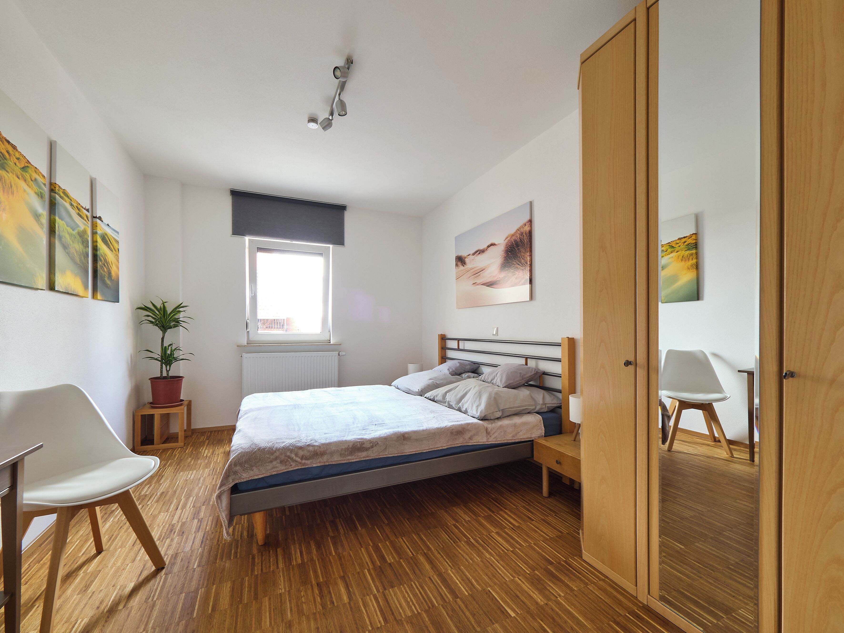 A bedroom with a bed, a window, a radiator, a potted plant, a chair, and a wooden wardrobe.