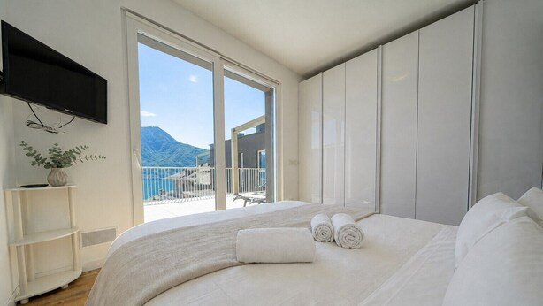 Ein Schlafzimmer mit einem großen Bett, einem an der Wand montierten Fernseher, einer Pflanze auf einem Regal und einem Balkon mit Bergblick.
