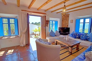 Ein Zimmer mit Holzbalkendecke, einer Couch, einem Fernseher und einem Fenster mit blauen Rollläden.