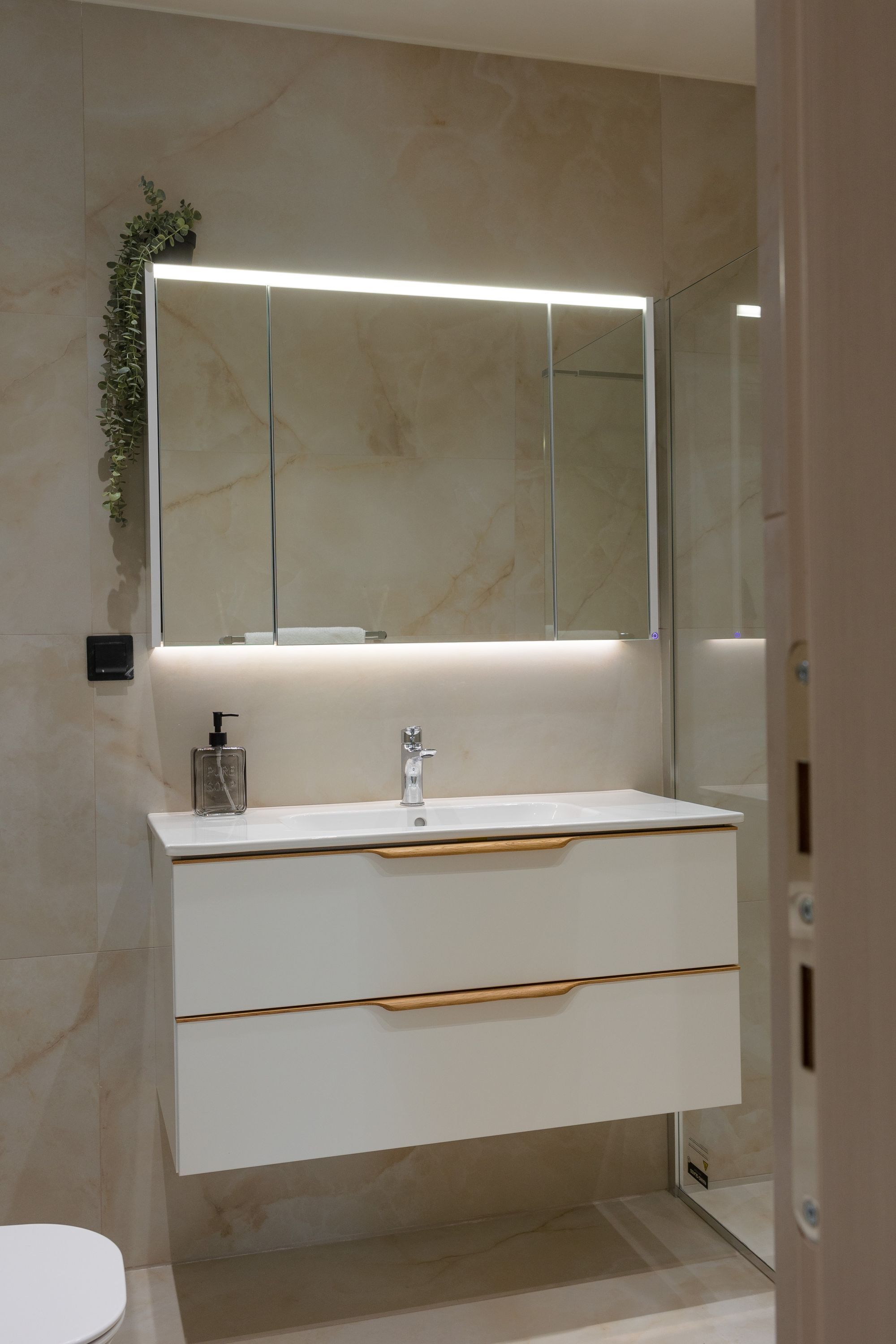 Een moderne badkamer met een marmeren wand, een wit wastafelmeubel met een spiegel van glas, en één lade.