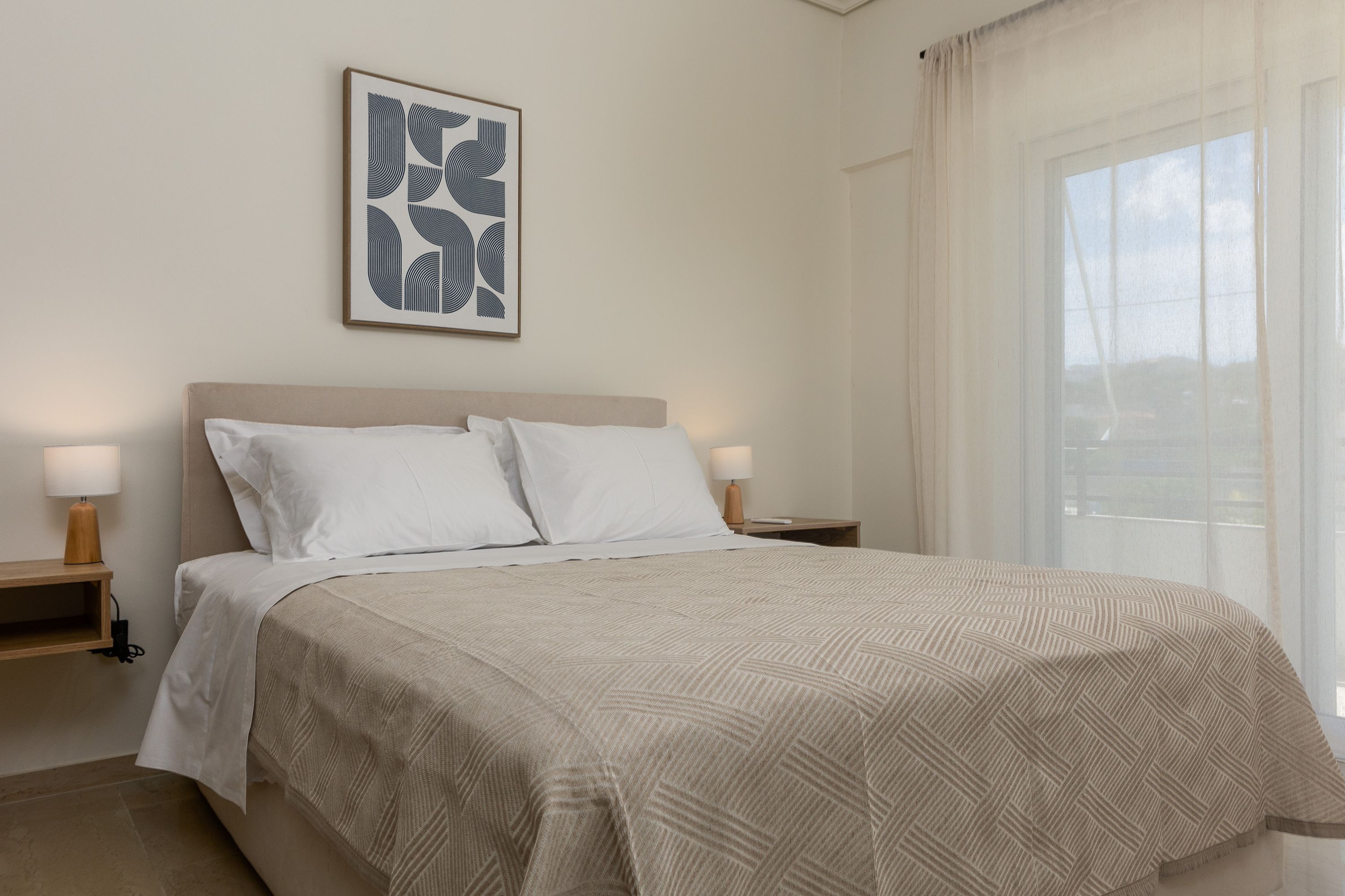 A neatly made bed with a geometric patterned bedspread, two bedside lamps, a framed abstract artwork on the wall, and a large window with sheer curtains.
