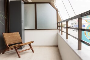 Un balcon avec un fauteuil en osier, une fenêtre avec des stores et une vue sur une rue où se trouve un immeuble bleu.