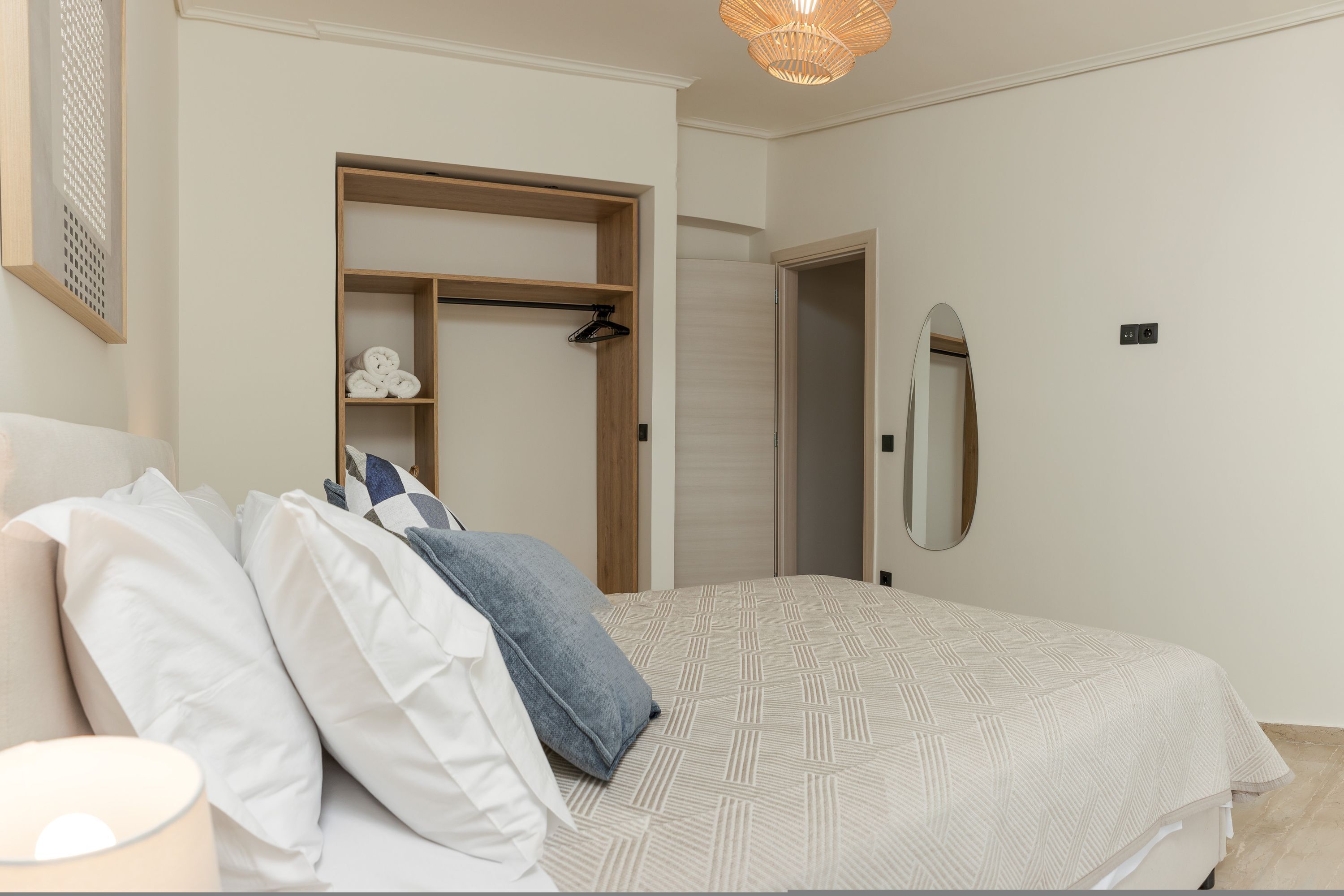 Een slaapkamer met een bed, kussens, een bedlampje, een houten garderobe, een spiegel en een hangende lamp.