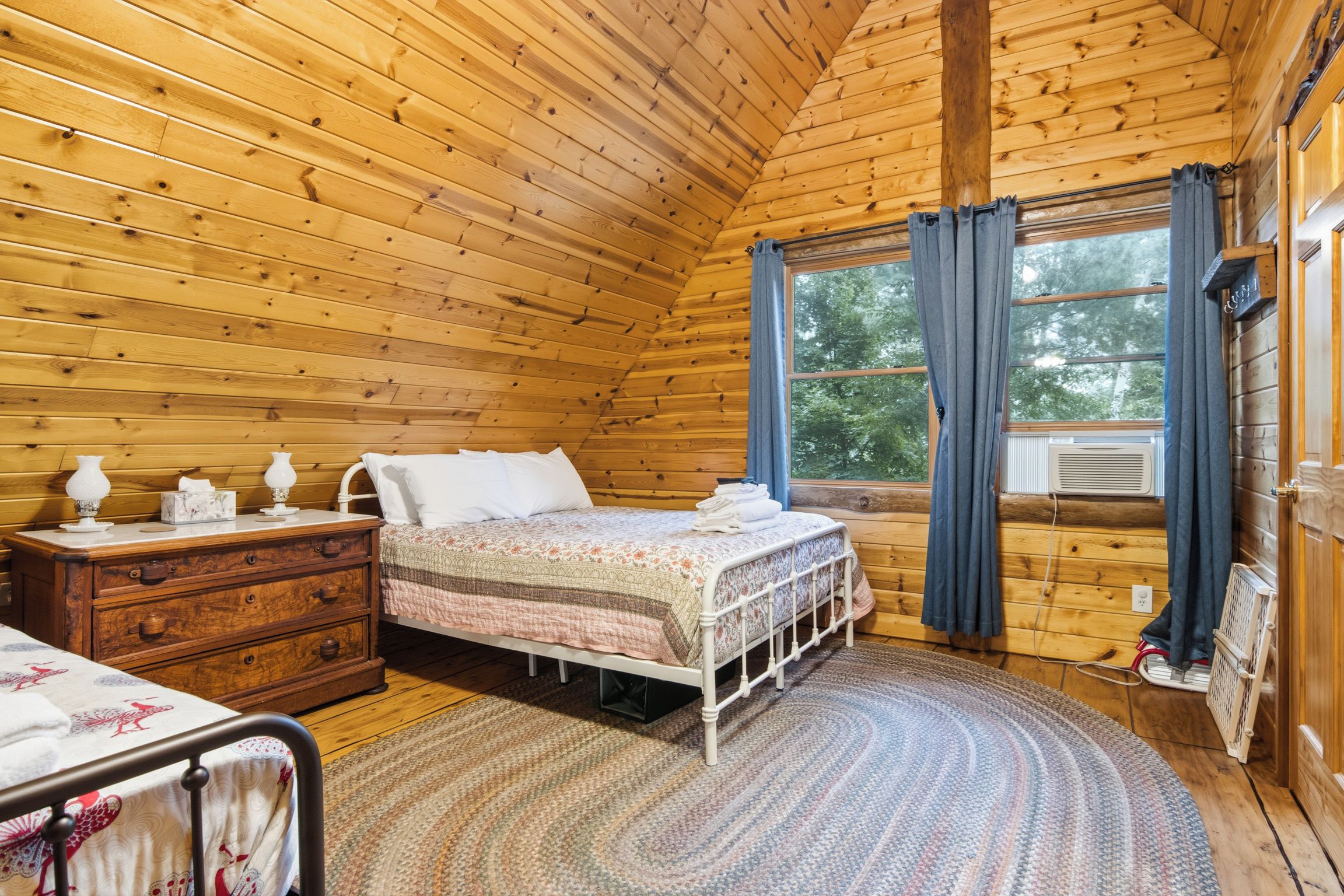 A wooden cabin bedroom with a bed, dresser, and window with curtains.