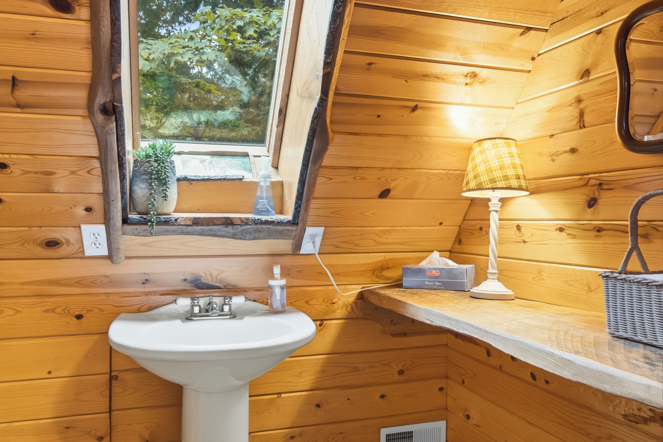 A wooden cabin bathroom with a window, a sink, a lamp, and a tissue box.