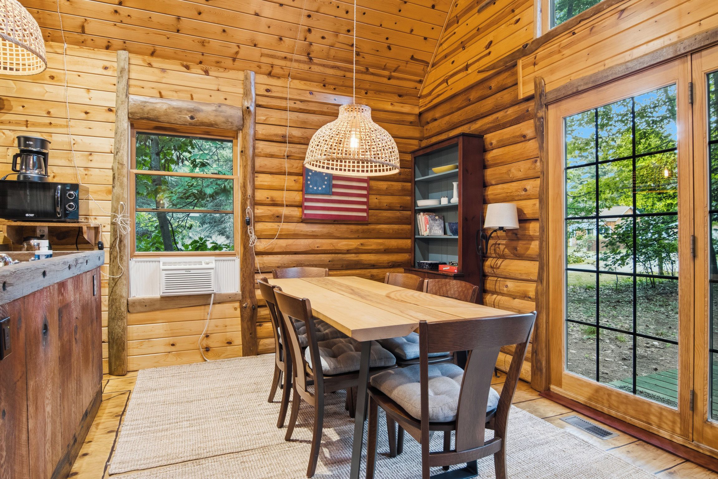 A wooden cabin interior with a dining table, chairs, a hanging light fixture, a coffee maker, and a bookshelf.