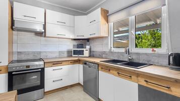A modern kitchen with white cabinets, a stainless steel oven, a dishwasher, a microwave, and a window with a view of greenery.