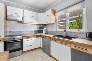 A modern kitchen with white cabinets, a stainless steel oven, a dishwasher, a microwave, and a window with a view of greenery. - Elena's House in Mournies (Μουρνιές)