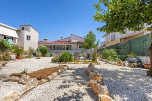 A residential area with a house featuring a red-tiled roof, a covered patio, and a garden with various plants and a gravel driveway. - Elena's House in Mournies (Μουρνιές)