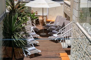 A sunlit outdoor lounge area with wooden flooring, white lounge chairs, a large umbrella, and potted plants. - Villa Constantia Armeni Rethymno (Armeni)