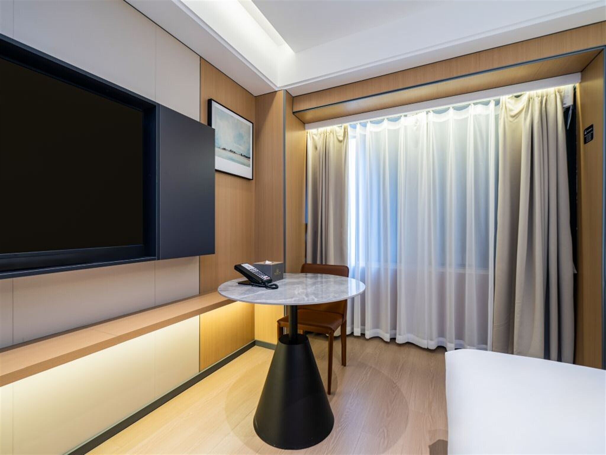 A hotel room with a flat-screen TV, a round table with a remote, a bed, and a window with curtains.