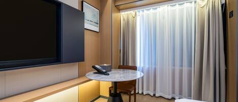 A hotel room with a flat-screen TV, a round table with a remote, a bed, and a window with curtains.